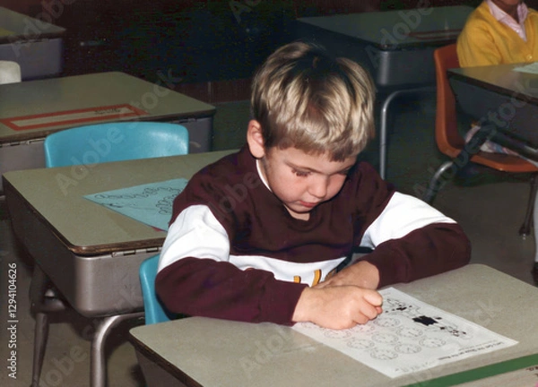 Fototapeta Circa 1987 - Minnesota: Student (boy) in first grade learns to tell time, sitting at his desk in elementary school classroom. Vintage scan, may have imperfections