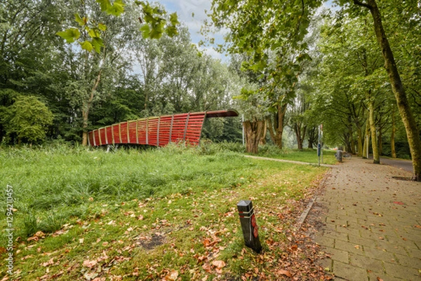 Obraz A striking modern red bridge blends seamlessly into a lush green park, surrounded by trees and leafy pathways, inviting exploration and relaxation.