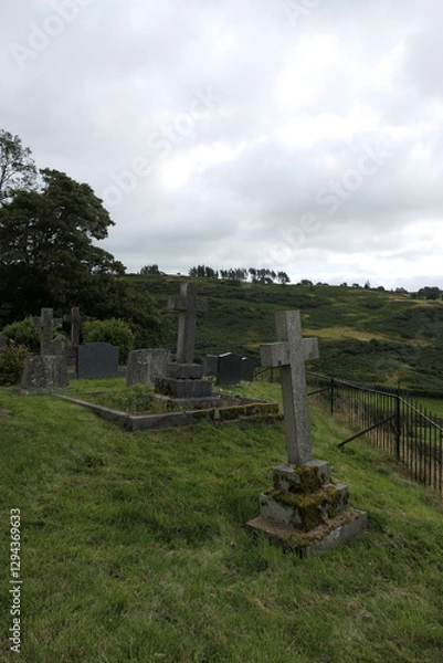 Fototapeta An old English graveyard with weathered tombstones covered in moss, standing amidst lush green grass. Some stones lean slightly, worn by time.