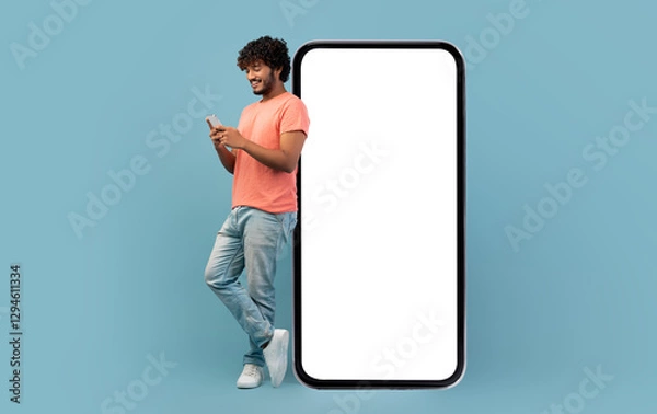 Fototapeta Cool curly indian guy in casual outfit leaning on huge smartphone with white blank screen, using cell phone and smiling, blue studio background, mockup, panorama with copy space