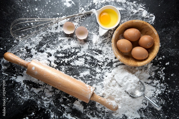 Fototapeta Baking cake ingredients. Bowl, flour, eggs, egg whites foam, egg beater, rolling pin and eggshells on black chalkboard