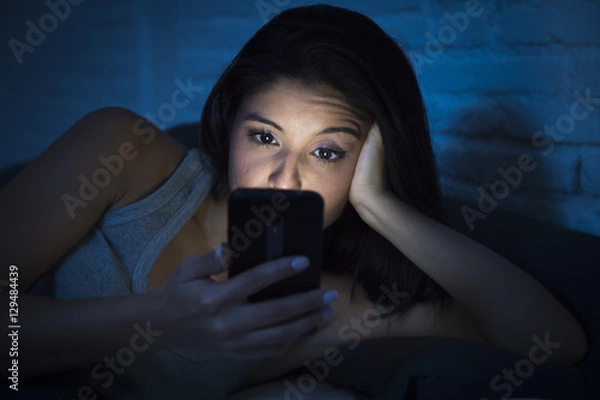 Fototapeta girl in bed using mobile phone late at night at dark bedroom lying happy and relaxed