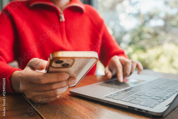 Fototapeta Close-up of a woman multitasking with a smartphone and laptop, wearing a red sweater, at a wooden table.