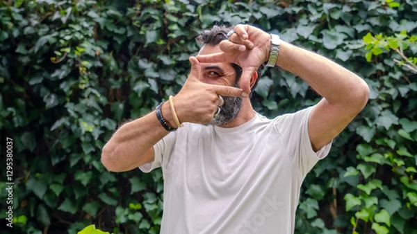 Obraz A middle-aged man with fair skin and a beard, standing outdoors in a wooded area, looking at the camera and bringing his hand close to his eye, making a camera lens gesture