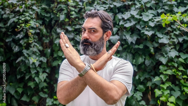 Obraz A middle-aged man with fair skin and a beard, outdoors in a wooded area, making a "no" gesture with his hands by crossing them, while looking at the camera