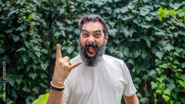 Obraz A middle-aged, fair-skinned, bearded, dark-haired man making the metal music hand gesture while looking at the camera in a wooded outdoor setting