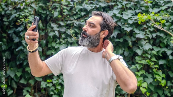 Obraz A middle-aged, fair-skinned, bearded, dark-haired man taking a selfie with his cellphone in a wooded area, in a close-up shot