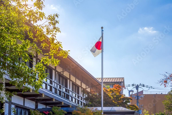 Fototapeta A Japanese flag waves atop a flagpole beside a traditional-style building, surrounded by lush greenery under a bright sky.