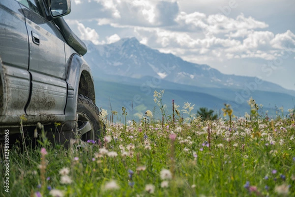 Fototapeta An off-road car on mud tires stands in a field with flowers against a background of snowy mountains and forests. The travel car is parked in a meadow. Tires for an SUV on the background of mountains.