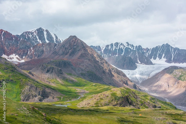 Fototapeta Vivid dramatic alpine scenery with green hills and rocks with view to high rocky pointy peak, sharp rockies, large snow-capped mountain top, snowy range and big glacier tongue under gray cloudy sky.