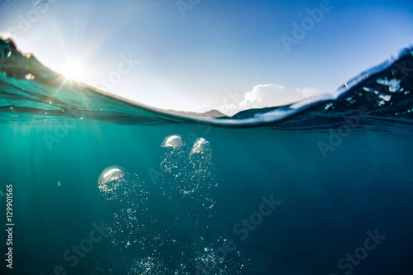 Fototapeta Ocean water line splits sky and underwaer part. Air bubbles in deep blue. Bright sun light and blue clear sky