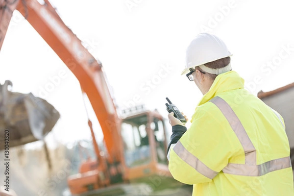 Fototapeta Supervisor using walkie-talkie at construction site against clear sky