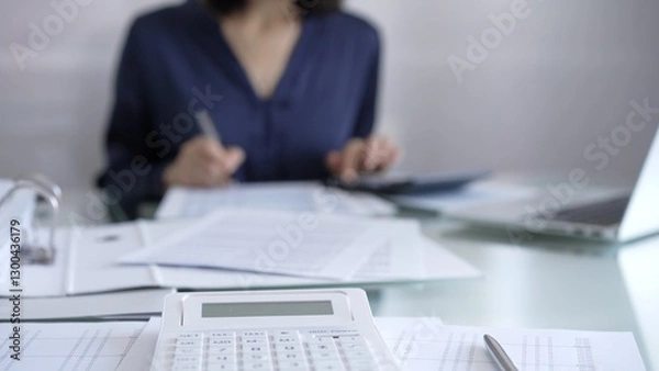 Fototapeta White calculator, pen and documents are lying on accountant's desk. Businesswoman wearing blue dress is working with documents on the background. Business and audit concept