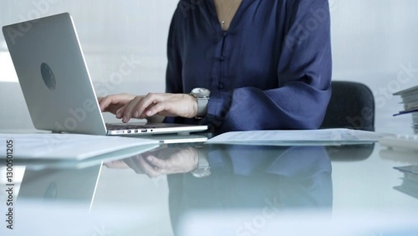 Fototapeta Businesswoman in blue blouse typing on laptop at glass desk while sitting in bright office setting, close-up vie. Businesspeople concept