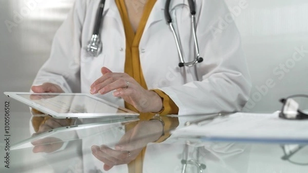 Fototapeta Female doctor, wearing yellow blouse under a medical coat, is using digital tablet in modern healthcare setting, interacting with patient data, close-up. Medicine concept