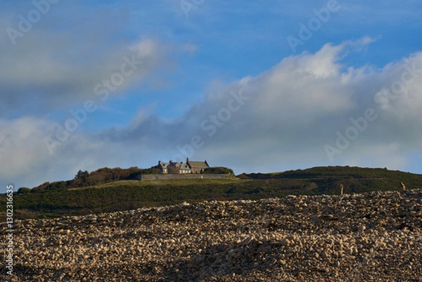 Fototapeta Scenic view of a traditional stone house atop a green hill in Normandy, France, with rocky terrain in the foreground and a clear blue sky with scattered clouds.