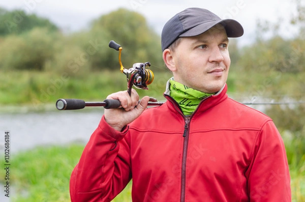 Obraz Fisherman in red sweatshirt and cap on the river bank with spinning rod in his hands.