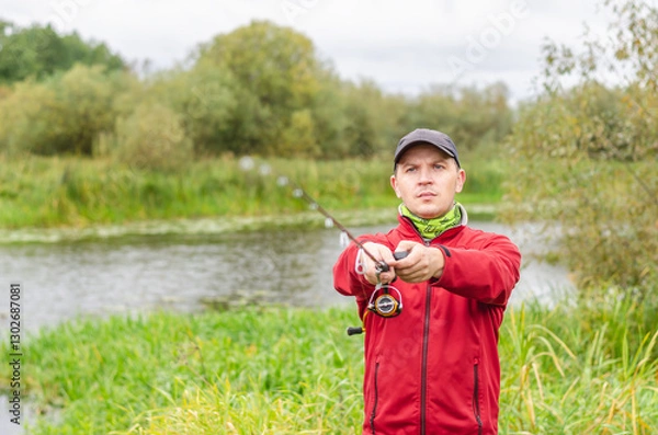 Obraz Fisherman in red sweatshirt and cap on the river bank with spinning rod in his hands.