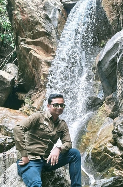 Fototapeta A man posing confidently in front of a stunning waterfall, surrounded by rugged rocks and lush greenery. The cascading water adds a serene and adventurous touch, capturing the beauty of nature.