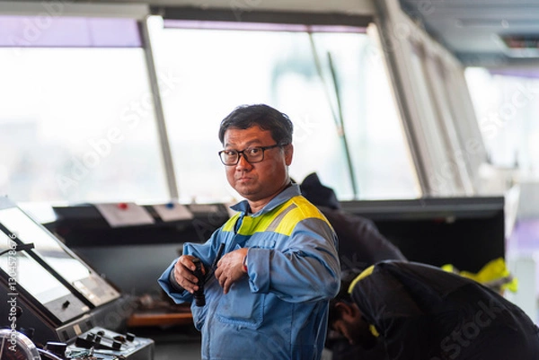 Obraz A marine engineer stands on the bridge of a large container ship, performing maintenance on navigation and communication equipment while the vessel is docked in port.