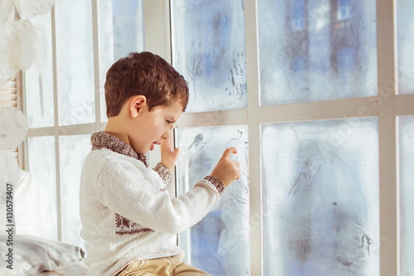 Fototapeta Portrait of little boy draws on a frozen window in the winter