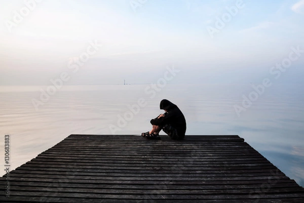 Fototapeta Frustrated depression man in the hood sitting on wooden bridge, near the beach on sunset. Concept of unemployed, sadness, depressed and human problems