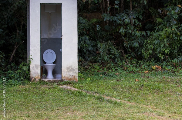 Fototapeta country house bathroom in the middle of a grassy area with the forest in the background with an open door and a view of a toilet with an open lid and a wall-mounted flush tank
