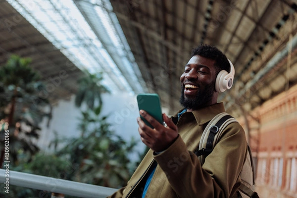 Obraz Tourist wearing headphones is using smart phone and smiling in airport or train station