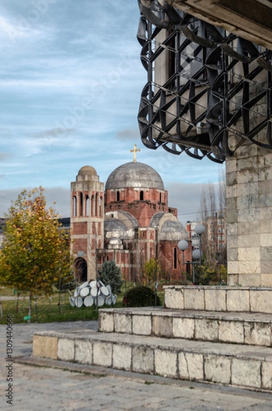 Fototapeta A partial view of the incomplete Christ the Saviour Cathedral in Pristina, Kosovo, framed by a modern architectural element. The cathedral stands against a partially cloudy sky