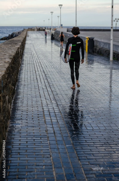 Fototapeta A barefoot surfer in a wetsuit walks along a wet promenade near the ocean, carrying a surfboard. The reflections on the wet pavement create a striking visual effect