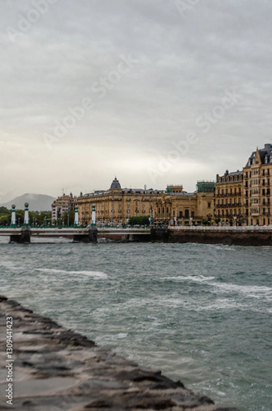 Fototapeta A serene view of San Sebastián and the Urumea River. The image captures the elegant architecture against a cloudy sky, with the river's gentle flow in the foreground
