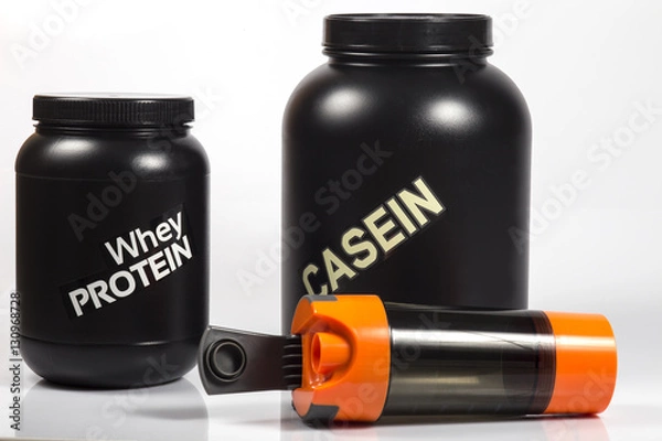 Fototapeta Whey protein black jar and big Casein protein black jar. Orange shaker lying near with opened cap. Close up image. White background. Shadows.