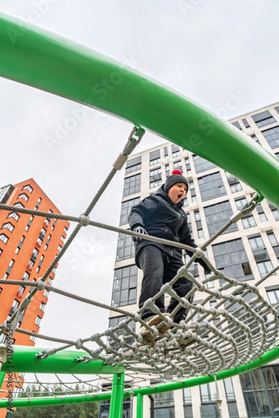 Fototapeta A child in warm clothes climbing a rope net on a playground surrounded by modern apartment buildings. Concept of outdoor activity in urban spaces