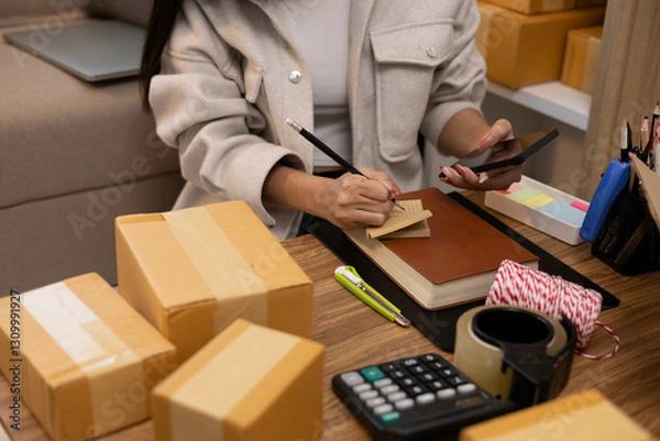 Fototapeta A woman is sitting at a table with a package in front of her. She is calculating expenses in a notebook and laptop and talking on her mobile phone.
