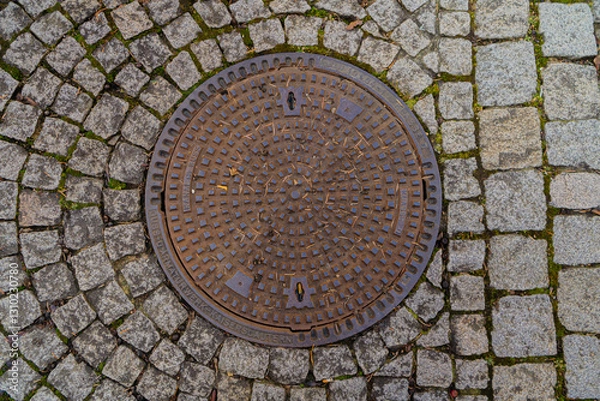 Obraz Round metal manhole cover on a cobblestone pavement. Urban detail, infrastructure. Close-up, top view