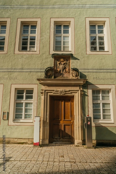 Obraz Building facade with a wooden door and decorative stone elements. Architectural detail, European architecture. "Das Diak." signage, educational center, sunlight and shadow.