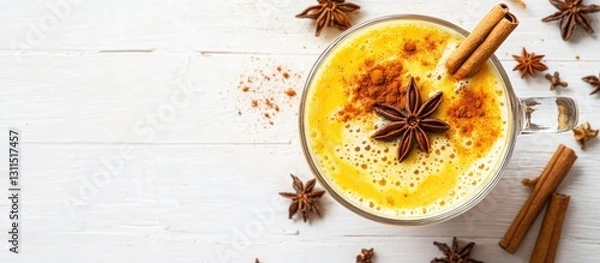 Fototapeta Golden turmeric latte in a glass topped with star anise and cinnamon sticks on a white wooden surface with spices around showcasing a warm vegan drink