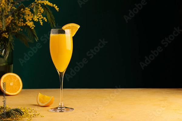 Fototapeta Mimosa cocktail in a champagne glass garnished with a triangle piece of orange. Alcoholic drink made from freshly squeezed orange juice and sparkling wine. Yellow table, dark background. Copy space 