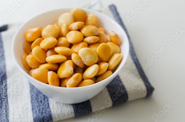 Fototapeta Lupin beans in a white bowl on a striped napkin on a white table. Italian food. Type of legumes. Healthy food concept. Horizontal orientation. Selective focus. Copy space.