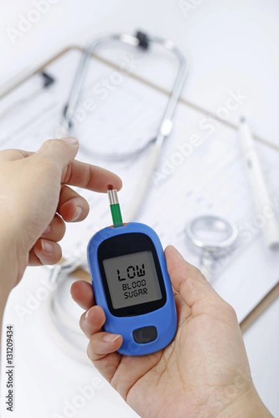Fototapeta Hand holding a blood glucose meter measuring blood sugar, the background is a stethoscope and chart file