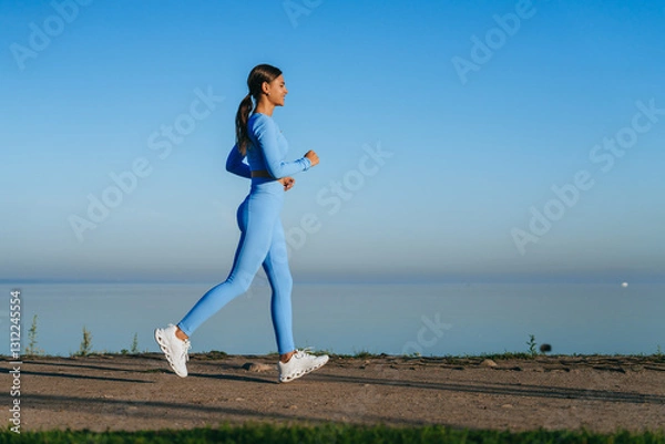 Fototapeta A woman in a vibrant blue outfit briskly walks along a serene waterside path, basking in the calmness of a clear morning sky.