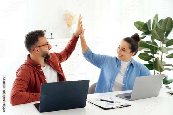 Fototapeta Cheerful a man and a woman high five while sitting at their desk in a modern office, celebrating a success together, both are working on their laptops and enjoying their time at work, smile