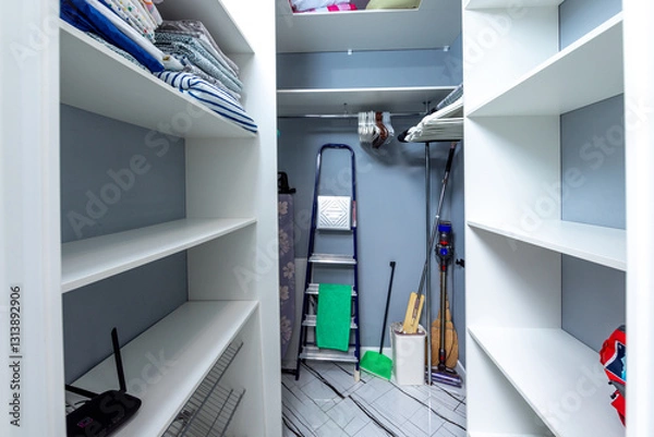 Obraz A small, clean, and organized closet with a ladder and a broom leaning against the wall. The closet is filled with clothes and other items, but it is well-organized and tidy