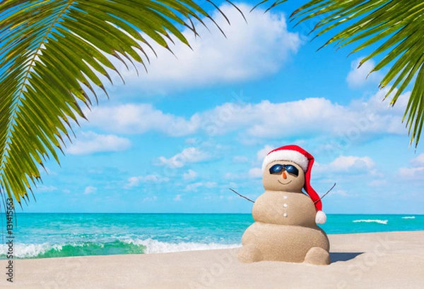 Fototapeta Sandy Snowman w świątecznym Santa kapeluszu i okularach przeciwsłonecznych na tropikalnej plaży oceanu