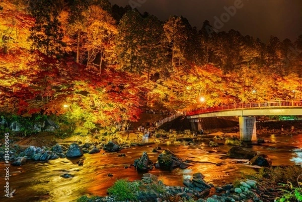 Fototapeta Korankei, Japan, glows with illuminated autumn foliage at night, highlighting a red bridge over a rocky river. Warm light reflects in the flowing water.