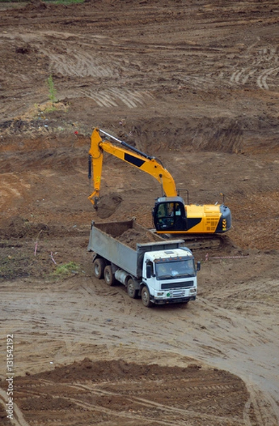 Obraz Рытьё котлована. Экскаватор нагружает грузовик-самосвал вырытым грунтом