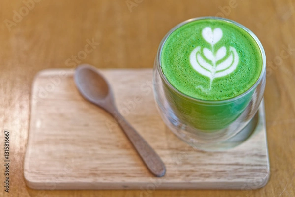 Fototapeta Matcha Latte with Beautiful Creamy White Foam Latte Art on a Wooden Tray. A freshly made green matcha latte served in a double walled glass cup, featuring a delicate latte art design on top.
