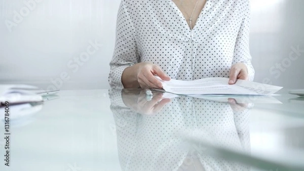 Fototapeta Businesswoman in white dotted blouse is going through paperwork while holding documents at a glass desk in a well lit, contemporary office setting. Business people concept