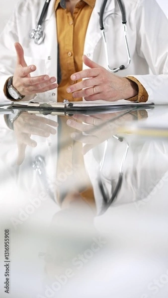 Fototapeta Unknown male doctor wearing a stethoscope, yellow shirt under a lab coat is gesturing with hands overs a glass desk with a laptop in clinic office. Medicine concept
