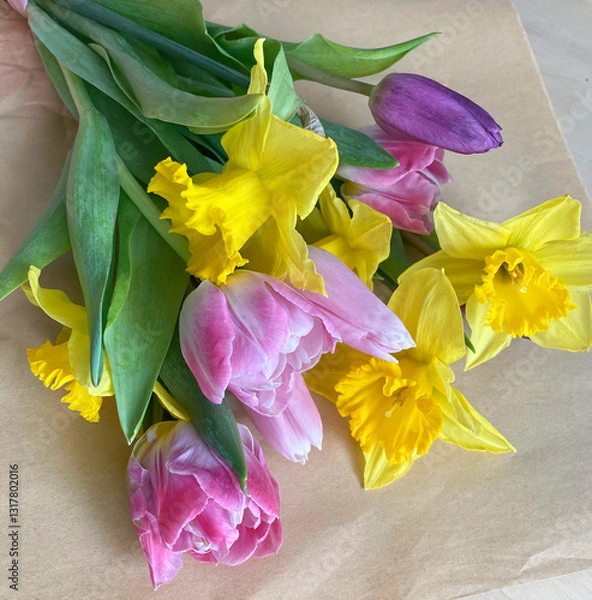 Fototapeta Beautiful composition of the first spring flowers with green leaves in a bouquet. Yellow daffodils, pink tulips in a bouquet on craft paper, top view.